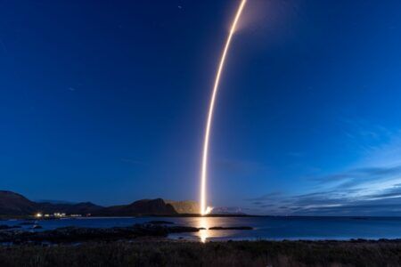 Alt text: A long-exposure night photograph of the Spectrum rocket rising from the Andøya coast, its bright arcing exhaust trail reflected in the water below against a deep blue twilight sky