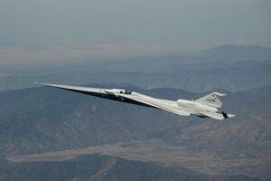 NASA’s X-59 quiet supersonic experimental aircraft banking in flight above mountainous desert terrain, its long pointed nose extending to the left and the NASA logo visible on the fuselage