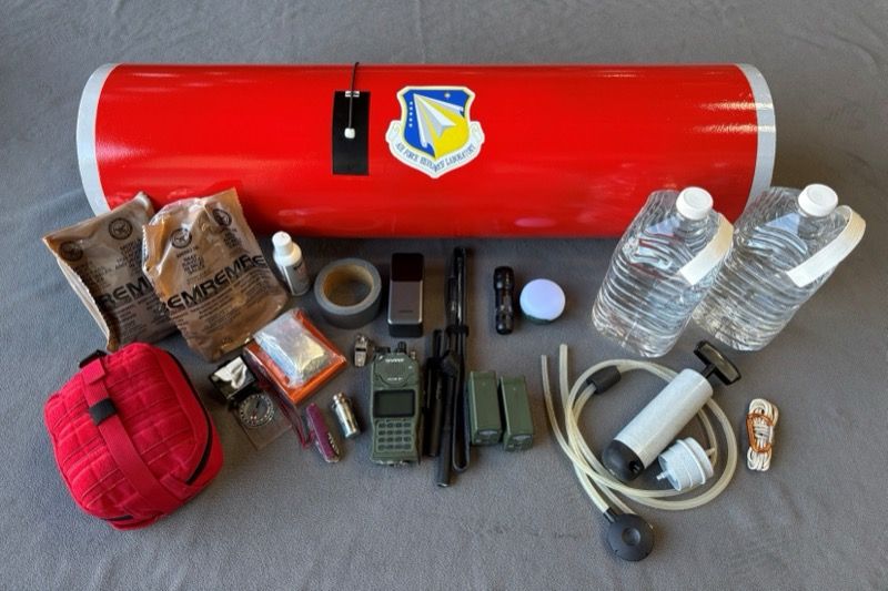 Survival and mission equipment laid out beside a red cargo pod, including ration packs, water bottles, a satellite radio, a desalinator pump, a life-raft bag and medical and signalling gear