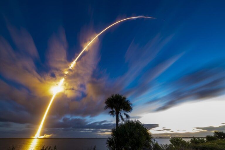 A long-exposure image of a rocket arcing into a dusk sky over water, leaving a bright trail above a silhouetted palm tree