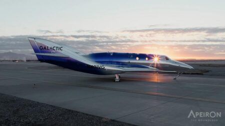 Virgin Galactic flies new Apeiron spaceplane to Spaceport America Silver and blue spaceship with GALACTIC lettering on its tail parked on an apron at sunrise, with the sun flaring through its cabin windows