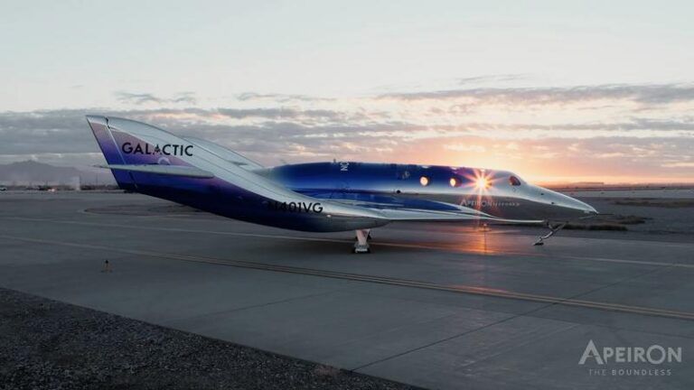 Virgin Galactic flies new Apeiron spaceplane to Spaceport America Silver and blue spaceship with GALACTIC lettering on its tail parked on an apron at sunrise, with the sun flaring through its cabin windows