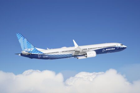 A Boeing 737-10 in Boeing house livery climbing against a blue sky above cloud, with “737” on the tail and a LEAP engine visible