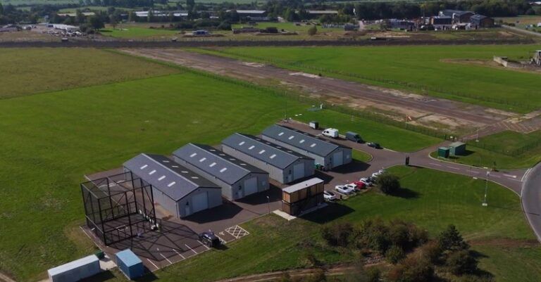 UK expands national drone testing center with new capabilities Aerial view of the drone testing facility at Westcott Venture Park, with hangars, a control building, parking and a test area beside a runway in open countryside