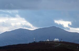 Radar domes and antennas of a weapons test range on a hilltop, with mountains and a cloudy sky behind