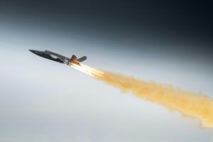 Dark grey uncrewed combat aircraft climbing steeply with a bright rocket booster flame and a long orange smoke trail against a grey sky