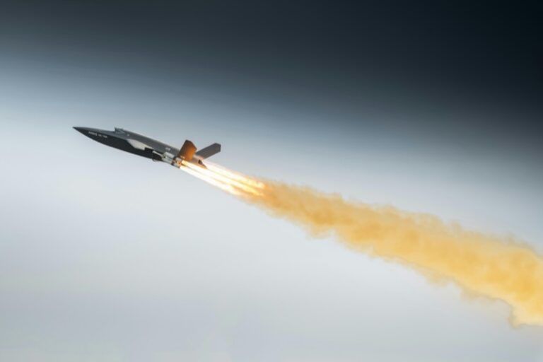 Dark grey uncrewed combat aircraft climbing steeply with a bright rocket booster flame and a long orange smoke trail against a grey sky