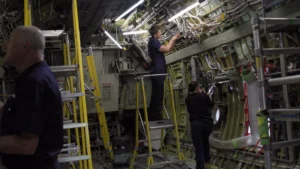 Engineers working inside an aircraft fuselage, one on a platform ladder routing bundles of electrical wiring overhead while others work on structures and systems nearby