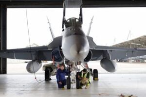Two technicians kneel to work on the nose landing gear of an F/A-18 fighter jet in a hangar, with the aircraft’s canopy raised