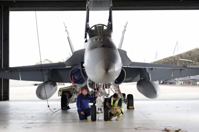 Australian project aims to certify laser repairs for fighter jets Two technicians kneel to work on the nose landing gear of an F/A-18 fighter jet in a hangar, with the aircraft’s canopy raised
